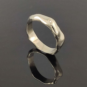 Op de afbeelding: Een zilverkleurige metalen ring met een onregelmatig, organisch en golvend bandontwerp. Het oppervlak van de ring is glad en sterk reflecterend, waardoor het licht heldere accenten creëert langs de ongelijkmatige randen van de buitenkant.