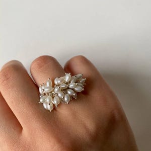 Op de afbeelding: Een zilveren ring met een cluster van kleine witte parels. De parels zijn in een losse, organische vorm gerangschikt, wat een delicate en vrouwelijke uitstraling creëert.