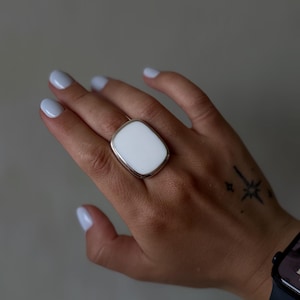 May include: A large statement ring featuring a smooth, rectangular white stone with rounded corners, set in a polished silver-toned metal band. The ring has a substantial, flat-faced design that sits prominently on the finger with a clean finish.