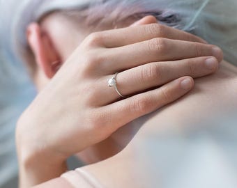 May include: A silver ring with a single pearl set in the center. The ring is worn on the finger of a person with light skin.