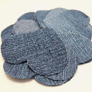 May include: A denim flower made from multiple layers of blue jean fabric. The flower has a layered petal design, with varying shades of blue. The fabric has a textured appearance.