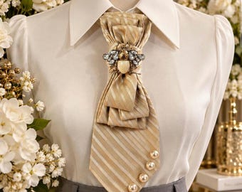 Può includere: Una camicia bianca con colletto abbinata a una cravatta beige a righe con una spilla decorativa a forma di ape con cristalli e un corpo di perla. La cravatta è decorata con quattro perle bianche rotonde sul bordo inferiore, sopra pantaloni grigi.