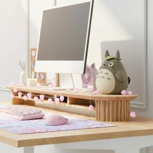 May include: A light wood desk with a computer monitor, keyboard, and mouse. A plush Totoro figure sits on a wooden monitor stand, adorned with a string of pink heart-shaped lights. A pink mousepad and fan complete the setup.