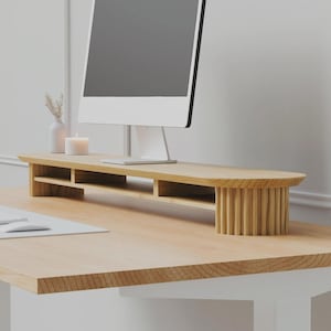 May include: A light-colored wooden monitor stand featuring a flat top surface, three open storage compartments underneath, and two rounded side supports with a vertical fluted texture. The stand is positioned on a desk holding a computer monitor.