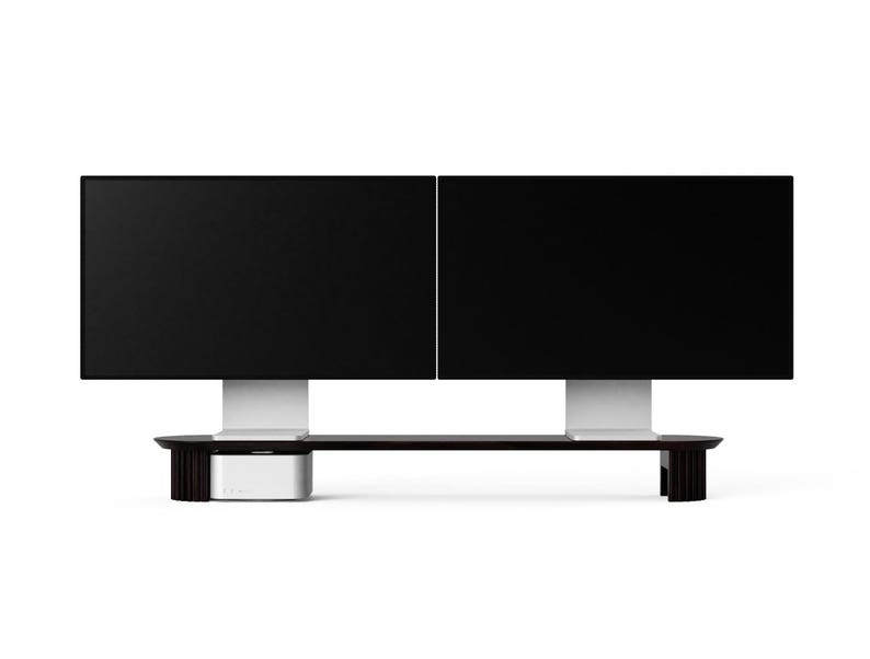 May include: A black monitor stand with a dark wood finish and a white shelf. Two black computer monitors are mounted on the stand.