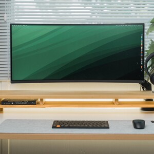 May include: A contemporary desk arrangement with a large computer monitor displaying a green gradient. A wooden monitor stand supports the screen, alongside a keyboard, mouse, and headphones. The desk is white, with a window and a plant in the background.
