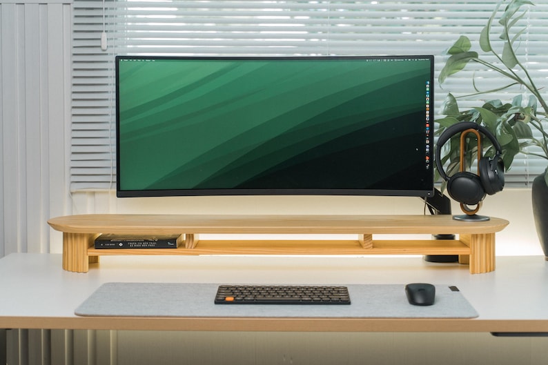 May include: A contemporary desk arrangement with a large computer monitor displaying a green gradient. A wooden monitor stand supports the screen, alongside a keyboard, mouse, and headphones. The desk is white, with a window and a plant in the background.