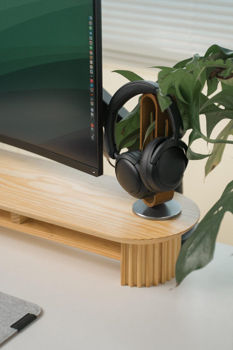 May include: A light wood desk with a monitor and black headphones. The desk features a rounded edge and a wooden headphone stand. The headphones are black. A green plant is in the background. The desk is approximately 100 cm wide.