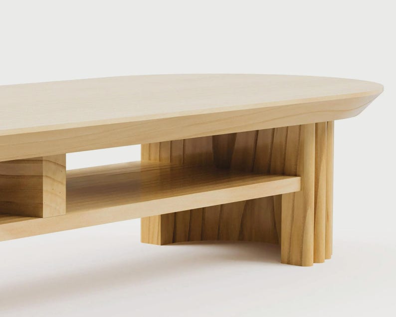 K&ouml;nnte beinhalten: Ein heller Couchtisch aus Holz mit einer glatten, ovalen Tischplatte und einer unteren Ablage. Der Tisch verf&uuml;gt &uuml;ber dicke, vertikal geriffelte Beine, die f&uuml;r strukturelle Stabilit&auml;t sorgen und die nat&uuml;rliche Maserung des Holzes zeigen.