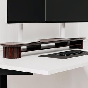 May include: A dark wood monitor stand with two shelves and a curved front. The stand is on a white desk with a black keyboard and a white mouse.