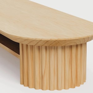 K&ouml;nnte beinhalten: Ein Couchtisch aus hellem Holz mit einer glatten, ovalen Tischplatte und einem zylindrischen Sockel mit vertikaler Rillenstruktur. Der Tisch weist eine sichtbare Holzmaserung sowie eine Ablagefl&auml;che unterhalb der Hauptoberfl&auml;che auf.