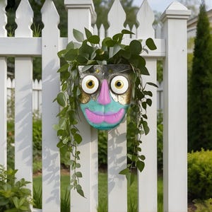 Op de afbeelding: Een grillige, gezichtsvormige plantenbak met een metallic afwerking, gouden accenten en een roze neus en lippen. De plantenbak is gevuld met groen gebladerte en hangt aan een witte hek, wat een speelse touch toevoegt aan de buitendecoratie.