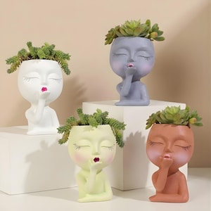 May include: Four decorative face planters in white, lavender, yellow-green, and terracotta colors, each holding a succulent plant. The planters have closed eyes, red lips, and are blowing a kiss. These unique planters are perfect for adding a touch of whimsy to any space.