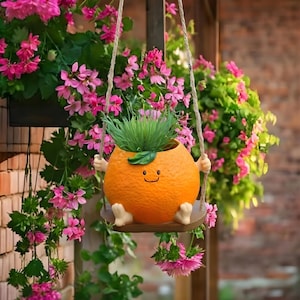 May include: Orange-shaped planter with a smiling face, arms, and feet, swinging from a wooden seat. The planter holds green grass and is suspended by rope. Pink flowers and greenery surround the planter, set against a brick wall.