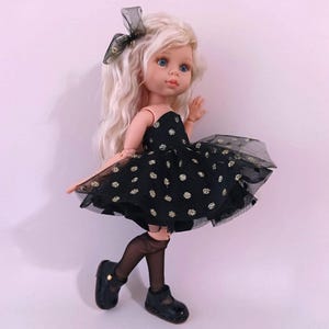 May include: A doll with long, wavy blonde hair and blue eyes, wearing a black dress with gold polka dots and a sheer overlay. The doll has black shoes and a matching bow. The doll is a toy.