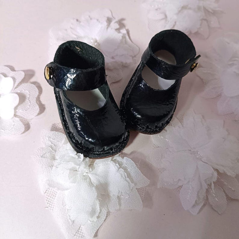 May include: A pair of small, black patent leather Mary Jane shoes. Each shoe features a strap with a gold-coloured buckle. The shoes are likely for a doll or child. They are displayed on a light pink surface with white floral decorations.
