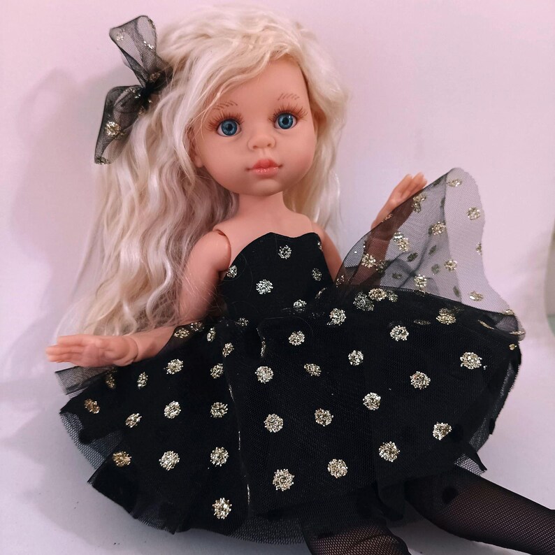 May include: A doll with long, wavy blonde hair and blue eyes. The doll is dressed in a black strapless dress with gold polka dots and a matching bow. The doll is wearing black tights.