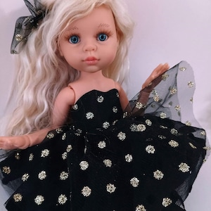 May include: A doll with long, wavy blonde hair and blue eyes. The doll is dressed in a black strapless dress with gold glitter polka dots and a matching bow. The doll has fair skin and a gentle facial expression.