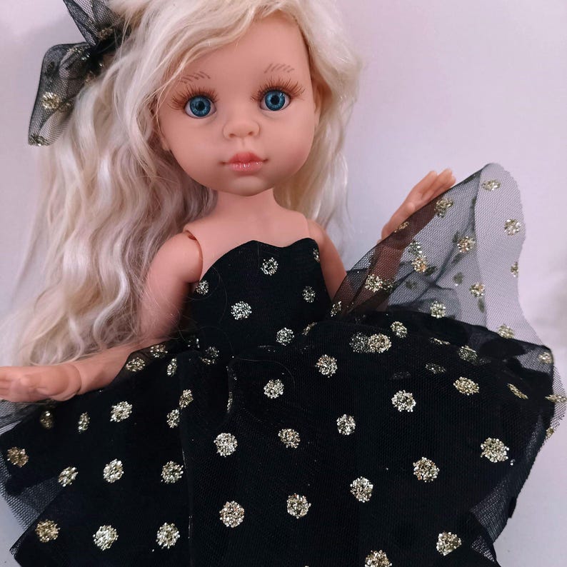 May include: A doll with long, wavy blonde hair and blue eyes. The doll is dressed in a black strapless dress with gold glitter polka dots and a matching bow. The doll has fair skin and a gentle facial expression.