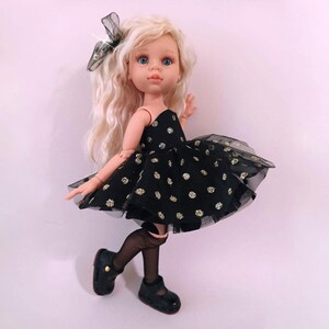 May include: A doll with long blonde hair, blue eyes, and a black dress with gold polka dots. The doll is wearing black shoes and stockings, and a black bow in her hair. The doll is posed against a light pink background.