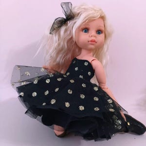 May include: A doll with blonde hair and blue eyes wearing a black dress with gold polka dots. The dress has a tulle overlay. A black bow with gold accents adorns the doll's hair. The doll is wearing black shoes.