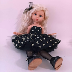 May include: A doll with long, wavy blonde hair and blue eyes. The doll is dressed in a black strapless dress with gold polka dots, black tights, and black shoes. A black bow with gold detailing is in the doll's hair.