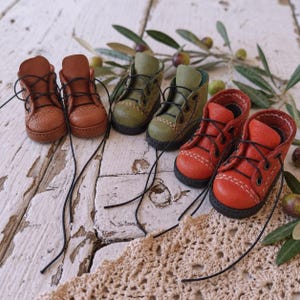 May include: Three pairs of miniature boots in tan, olive green, and orange. Each pair features black laces, metal eyelets, a rounded toe cap with visible white stitching, and a dark sole. The boots are arranged in a row on a white surface.