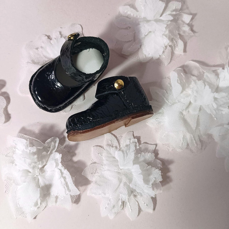 May include: Miniature black patent leather shoes, one Mary Jane style and one boot, with gold-coloured button fastenings. White lace flowers are scattered around the shoes.