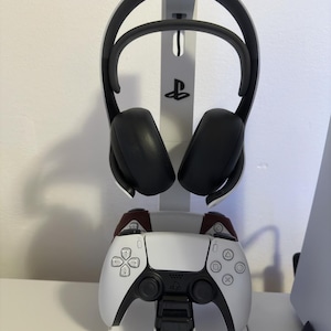 May include: A white and black gaming headset and controller stand. The stand holds a black and white controller with burgundy accents and a black headset. The stand has the PlayStation logo.