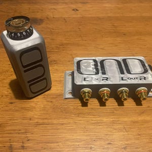 May include: A silver audio crossover box with the word "ONO" printed on the top. The box has four gold-coloured speaker terminals labelled "L IN", "R", "L OUT", and "R".