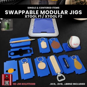 May include: A collection of blue modular jigs with various items, including keychains, a pen, and a baseball. The image includes the text "SWAPPABLE MODULAR JIGS" and "3D JIG SOLUTIONS".