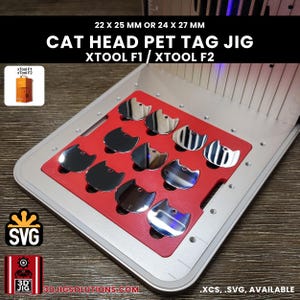 May include: A red cat head pet tag jig for xTool F1 and xTool F2 laser machines, featuring twelve slots for 22 x 25 mm or 24 x 27 mm tags. The jig is mounted on a silver metal base and includes text labels for compatibility and file formats.