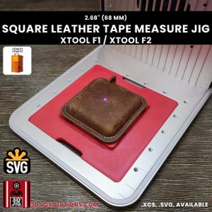 Könnte beinhalten: Eine quadratische Leder-Maßband-Vorrichtung für xTool F1 und xTool F2 Lasermaschinen. Die Vorrichtung verfügt über eine rote Grundplatte mit einem 68 mm quadratischen Ausschnitt, um ein braunes Leder-Maßbandgehäuse sicher zu fixieren.