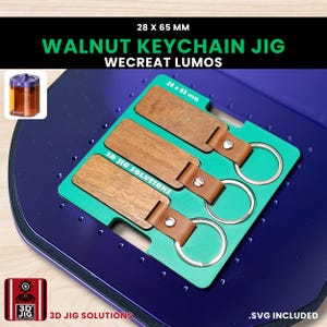 Könnte beinhalten: Eine Walnuss-Schlüsselanhänger-Jig mit drei Schlüsselanhängern, jeweils mit einem braunen Lederriemen und einem silbernen Ring. Die Jig ist grün und liegt auf einer blauen Oberfläche. Der Text "WALNUT KEYCHAIN JIG" und "3D JIG SOLUTIONS" sind sichtbar. Die Schlüsselanhänger messen 2,8 x 6,5 cm.