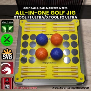 Könnte beinhalten: Eine gelbe Golfvorrichtung mit dem Text "ALL-IN-ONE GOLF JIG" und "XTOOL F1 ULTRA/XTOOL F2 ULTRA". Die Vorrichtung hält Golfbälle, Tees und Ballmarker. Es gibt zwei orangefarbene und zwei blaue Golfbälle sowie mehrere Holz-Tees.