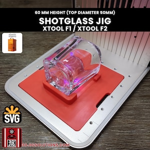 Puede incluir: Una plantilla de plástico rojo para vasos de chupito para máquinas láser xTool F1 y xTool F2, con 60 mm de altura y 50 mm de diámetro superior. La plantilla está montada sobre una base de metal plateado, con el texto SHOTGLASS JIG XTOOL F1 / XTOOL F2.