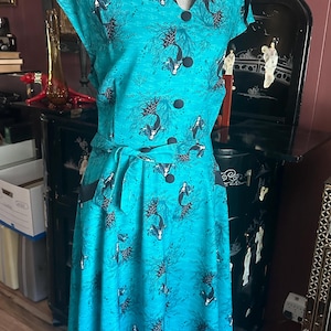 May include: A teal dress featuring a repeating pattern of black mermaids and seaweed. The dress has a V-neckline with black trim, short sleeves, five black buttons down the front, and a matching fabric tie belt at the waist with two side pockets.