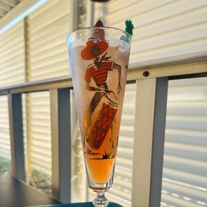 May include: A tall glass with a pink drink and a design of a person playing a drum. The glass has a clear stem and a round base.