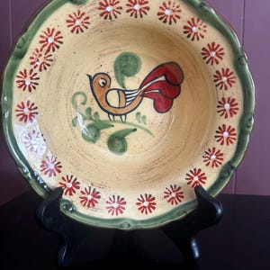 May include: A decorative ceramic bowl with a yellow interior and a green rim. The bowl features a painted bird design in orange, red, and green, surrounded by red and white floral accents. The bowl is displayed on a black stand.