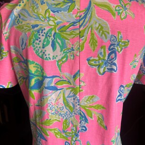Pode incluir: Uma blusa rosa de manga curta com um padr&atilde;o floral vibrante em tons de verde, azul e amarelo. O design apresenta flores e folhas estilizadas sobre um fundo rosa brilhante. A blusa tem decote em V e uma costura vertical nas costas.