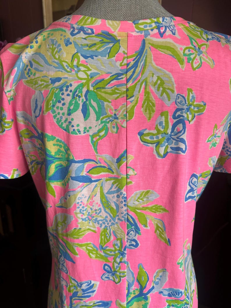 Pode incluir: Uma blusa rosa de manga curta com um padr&atilde;o floral vibrante em tons de verde, azul e amarelo. O design apresenta flores e folhas estilizadas sobre um fundo rosa brilhante. A blusa tem decote em V e uma costura vertical nas costas.