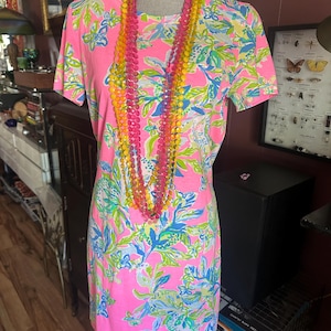 Pode incluir: Um vestido rosa de manga curta com um padr&atilde;o floral em azul, verde e branco. Um colar de contas multicolorido em tons de rosa, amarelo e laranja pende sobre o vestido. O vestido &eacute; exibido em um manequim.
