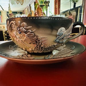 May include: A ceramic teacup and saucer set featuring a raised, three-dimensional dragon design. The cup and saucer have a gradient finish transitioning from dark charcoal at the rims to a lighter, muted beige tone, with detailed dragon scales.