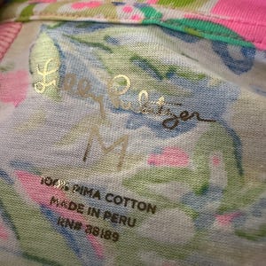 Pode incluir: Close-up de um tecido branco, rosa, azul e verde estampado com o texto dourado "Lilly Pulitzer" e o tamanho "M". A etiqueta diz "100% PIMA COTTON MADE IN PERU RN# 88189".