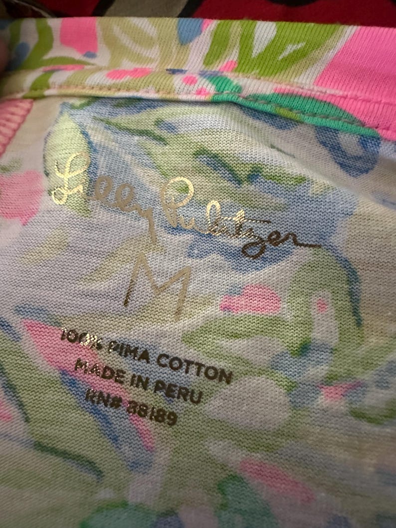 Pode incluir: Close-up de um tecido branco, rosa, azul e verde estampado com o texto dourado "Lilly Pulitzer" e o tamanho "M". A etiqueta diz "100% PIMA COTTON MADE IN PERU RN# 88189".