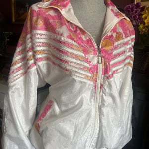 Op de afbeelding: Witte jas met bloemenprint en een volledige ritssluiting. De jas heeft een roze en geel bloemenpatroon op de schouders en de bovenrug. De jas heeft een witte basis met een getextureerde, gekreukelde uitstraling.