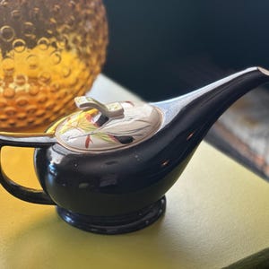 May include: A black ceramic teapot with a long spout and a handle. The lid is decorated with a floral pattern. A decorative glass orb is in the background.
