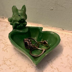 May include: A green ceramic heart-shaped trinket dish with a small bunny figurine on the side. The dish holds several rings. The rings are silver and gold, with one featuring a dark stone.