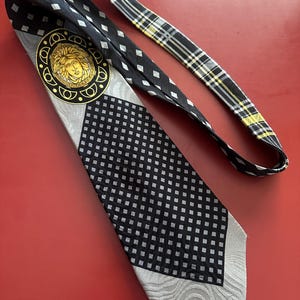 May include: A black and gray patterned necktie with a gold medallion featuring a face. The tie has a checkered pattern, a diamond pattern, and a plaid pattern on the back. The tie is laying on a red surface.