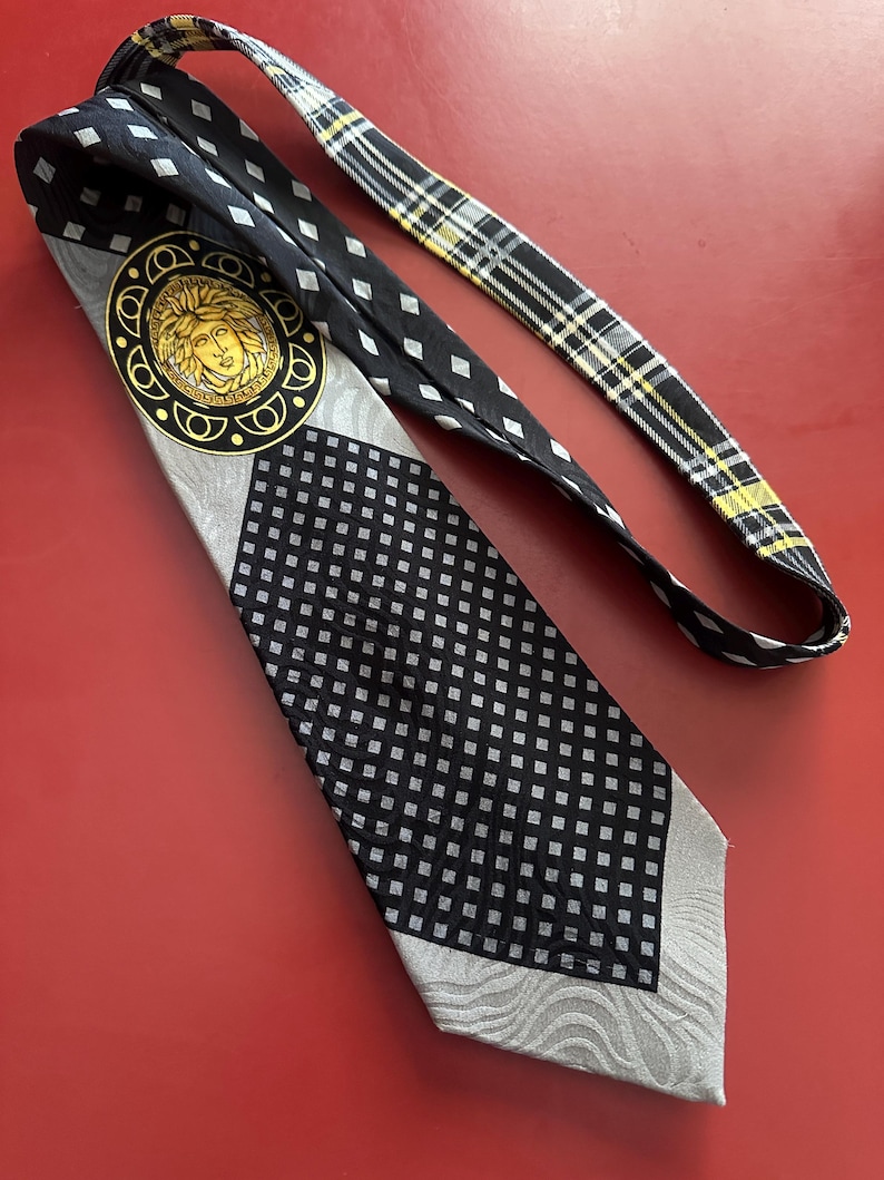 May include: A black and gray patterned necktie with a gold medallion featuring a face. The tie has a checkered pattern, a diamond pattern, and a plaid pattern on the back. The tie is laying on a red surface.